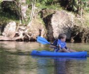 small boy in blue lifejacket on blue kayak with blue paddle on river in front of large rocks