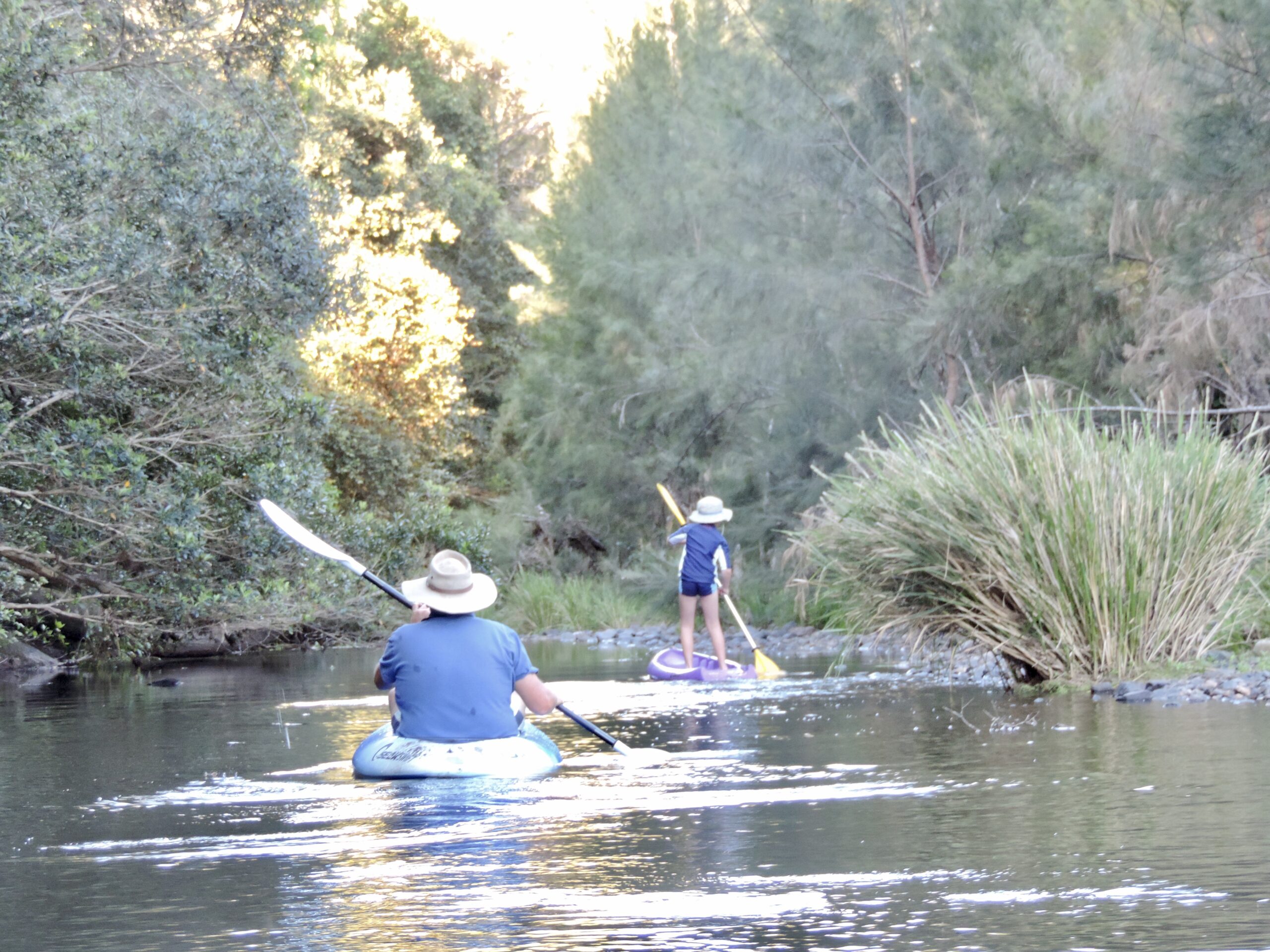 Man paddling kayak and child standing on small kayak with paddle in shallow Australian river