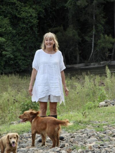 Blonde haired middle aged woman in long white tunic, shorts and wellington boots with two dogs at foreground